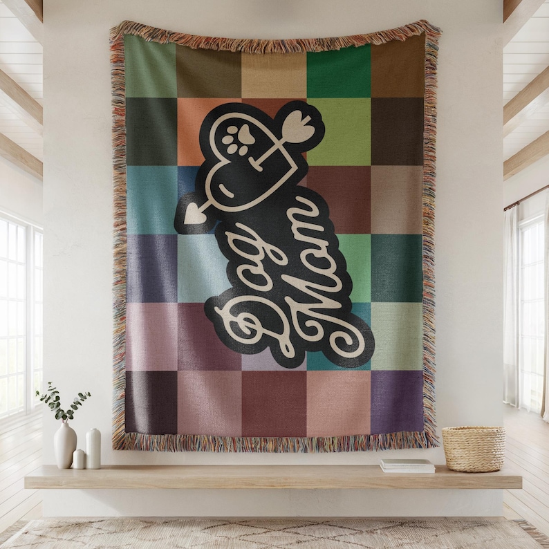 Op de afbeelding: Een geweven sprei met een patchwork-ontwerp in tinten groen, bruin, blauw en paars. De deken heeft de woorden "Dog Mom" in een cursief lettertype, met een hart en een pootafdruk. De deken heeft een gefranjerde rand.