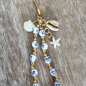 May include: A white and blue floral patterned phone charm with gold beads, a gold chain, a white seashell, and a white starfish.