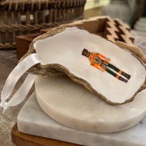 Pode incluir: Um ornamento de concha de ostra com uma figura de quebra-nozes em miniatura em laranja, preto e verde. A concha é presa a uma fita branca e repousa sobre uma pilha de bases de mármore e madeira. Um item decorativo de férias.