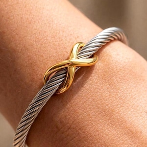 Könnte beinhalten: Nahaufnahme eines silberfarbenen Armbands mit gedrehtem Kabel und einem goldfarbenen Unendlichkeitsknoten. Das Armband hat ein detailliertes Design, das den Kontrast zwischen Silber und Gold hervorhebt. Ein modisches Schmuckstück.