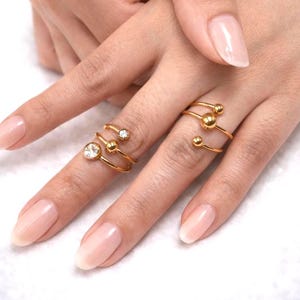Puede incluir: Anillos dorados en espiral con detalles esféricos y de cristal. Los anillos se muestran en una mano con uñas cuidadas. La joyería presenta un diseño moderno con una estética minimalista, adecuada para el uso diario o ocasiones especiales.