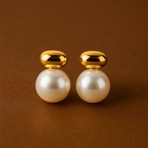 May include: A pair of elegant earrings featuring a large, creamy white pearl base topped with a small, gold-colored cap. The earrings are set against a brown background, highlighting their simple yet sophisticated design.