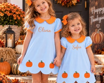 May include: Two light blue dresses with orange Peter Pan collars, orange sleeve cuffs, and scalloped hems. Each dress features embroidered orange pumpkins along the bottom and the names Olivia and Sofia embroidered in orange on the chest.