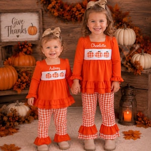 May include: Two matching orange long-sleeve shirts and orange and white gingham pants sets. Each shirt features a white chest panel with three embroidered pumpkins and a personalized name, Aubrey or Charlotte, in white script lettering.