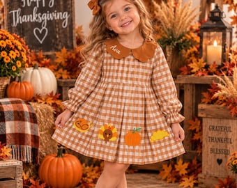 May include: A long-sleeved brown and white gingham dress with a scalloped collar embroidered with the name Ava. The dress features embroidered patches of a turkey, a pumpkin, and autumn leaves, paired with brown suede boots and a hair bow.