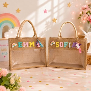 Puede incluir: Dos bolsos de yute rectangulares con paneles de plástico transparente en la parte frontal. Cada bolso presenta letras coloridas de estilo afelpado que deletrean los nombres EMMA y SOFIA. Un pequeño arcoíris y un unicornio cuelgan de cada asa.