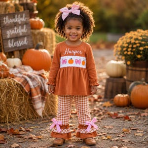 May include: A long-sleeve rust-colored tunic with a white chest panel featuring embroidered pumpkins and pink bows, personalized with the name Harper. The outfit includes matching rust and white gingham pants with ruffled hems and pink bows.