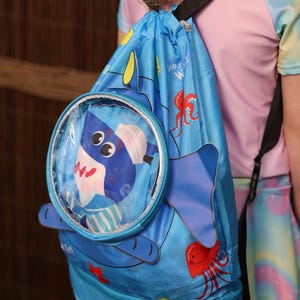 May include: Blue drawstring backpack with a shark design. The backpack features a clear circular pocket with a shark graphic, a zippered bottom compartment, and the name "Jason" printed on it. The bag also has illustrations of sea creatures.