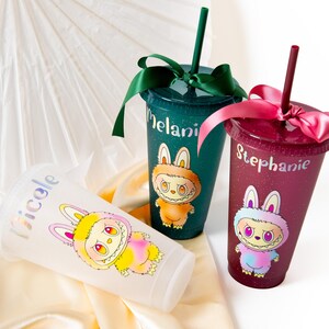 May include: Three personalized glitter tumblers with straws and bows. One is clear, one is teal, and one is burgundy. Each tumbler features a cartoon bunny character and a name: Nicole, Melani, and Stephanie.