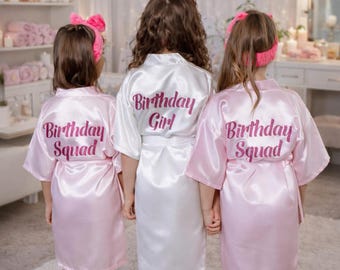 Puede incluir: Tres batas de satén con diademas a juego. Dos batas de color rosa claro llevan el texto Birthday Squad en rosa oscuro, mientras que la bata blanca central lleva el texto Birthday Girl en rosa oscuro. Mangas cortas y cinturón.