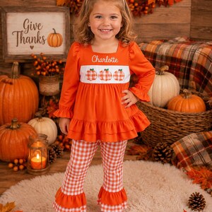 May include: A long-sleeved orange tunic with a white chest panel featuring three orange gingham pumpkins and the name Charlotte embroidered in white. The tunic has a ruffled hem and is paired with matching orange and white gingham bell-bottom pants.