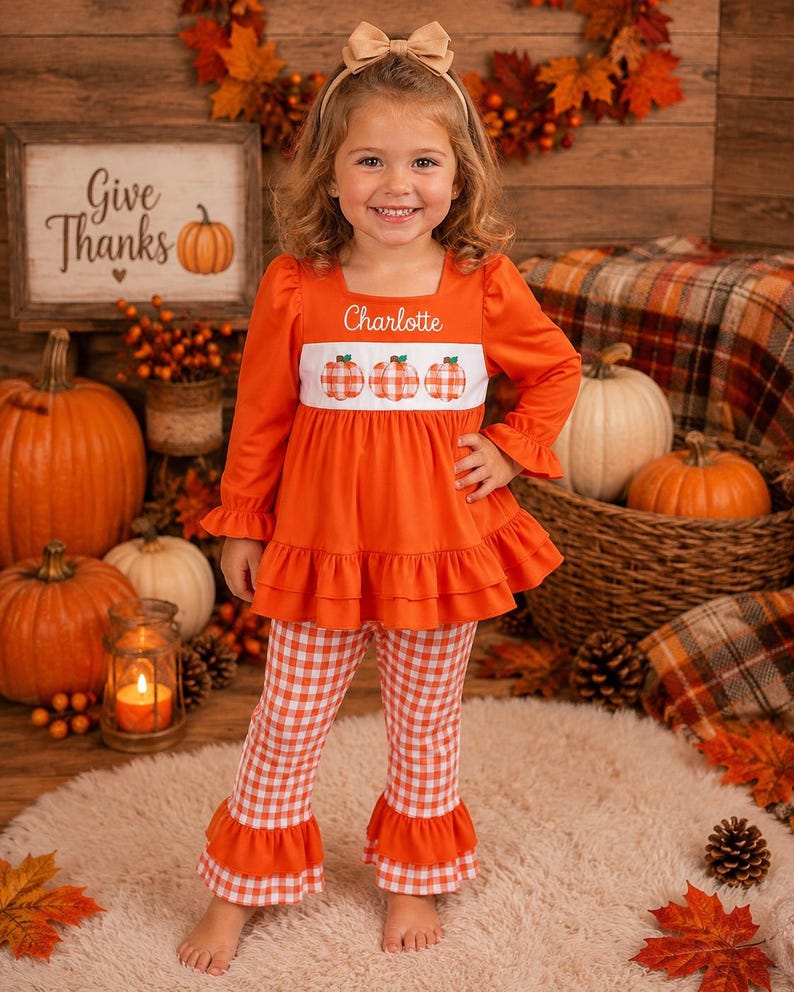 May include: A long-sleeved orange tunic with a white chest panel featuring three orange gingham pumpkins and the name Charlotte embroidered in white. The tunic has a ruffled hem and is paired with matching orange and white gingham bell-bottom pants.