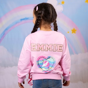 Peut inclure: Sweat-shirt rose avec le nom "EMMIE" en lettres dorées au dos. Un patch en sequins en forme de cœur avec un motif de licorne se trouve sous le nom.
