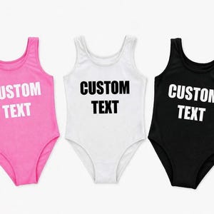 May include: Three custom text bodysuits in pink, white, and black. Each bodysuit has a scoop neck and is sleeveless. The words "CUSTOM TEXT" are printed in bold white letters on the pink and black bodysuits and in black on the white bodysuit.