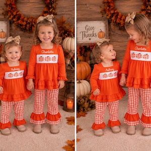 May include: Two matching orange long-sleeve tops with white smocked panels featuring embroidered pumpkins and the names Aubrey and Charlotte, paired with orange and white gingham print bell-bottom pants with ruffled hems and tan hair bows.