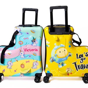 May include: Two colorful children's rolling suitcases. One is light blue, pink, and yellow with unicorns and donuts. The other is yellow with a cartoon astronaut and the text "Let's go Travel!" Both have black handles and wheels.