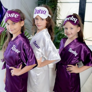 May include: Three girls wearing purple and white satin robes with matching eye masks. The eye masks and robes are personalized with the names "Rose", "Anna", and "Becky".