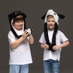 May include: Two children wearing black and white animal-themed hats with long ear-like flaps. The boy on the left is holding a small black and white plush toy. Both children are wearing white t-shirts and blue jeans.