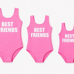 May include: Three matching pink swimsuits with the text "BEST FRIENDS" in white block letters. The swimsuits are in different sizes, suggesting a set for a group or family.