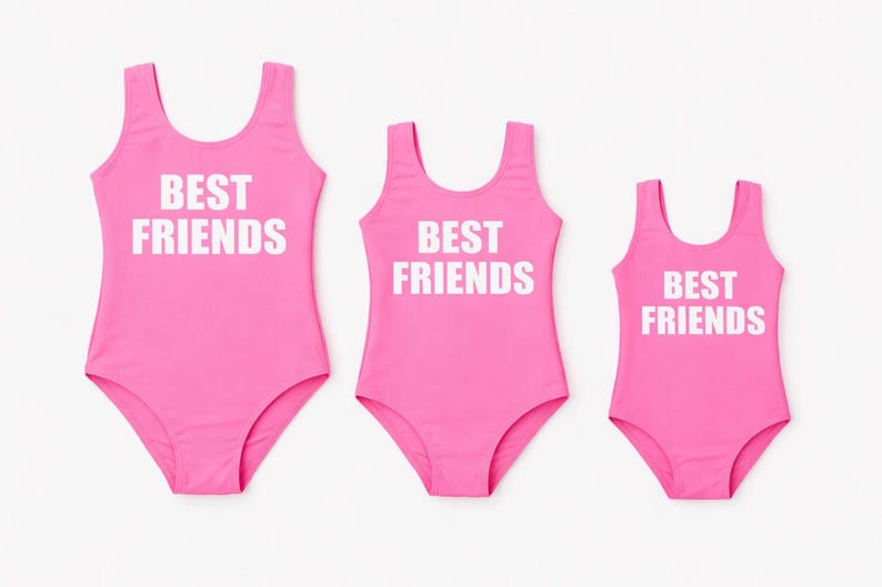 May include: Three matching pink swimsuits with the text "BEST FRIENDS" in white block letters. The swimsuits are in different sizes, suggesting a set for a group or family.