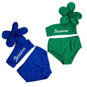 May include: Two girls' one-piece swimsuits with a flower detail on the shoulder strap. The swimsuit on the left is blue with the name "Jessica" embroidered on the top. The swimsuit on the right is green with the name "Jasmine" embroidered on the top.