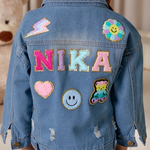 May include: A blue denim jacket with colorful patches, including a lightning bolt, a flower, a heart, a smiley face, a teddy bear, and the name "NIKA" in pink, purple, light blue, and pink letters.