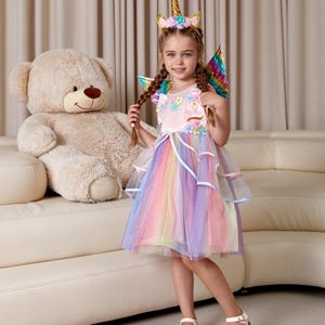 May include: A young girl wearing a pink unicorn dress with rainbow tulle and a unicorn headband. She is holding her braided hair and smiling. The dress has a floral design and the words "Sophia" embroidered on the bodice. She is standing in front of a white couch with a large teddy bear.