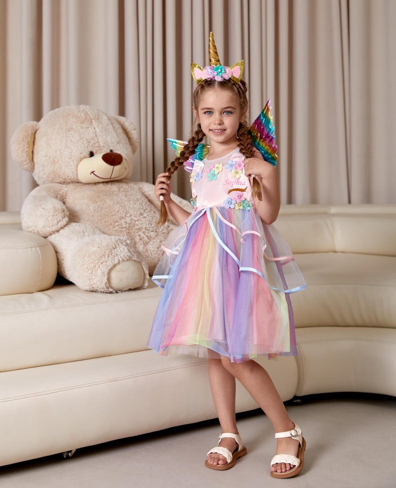 May include: A young girl wearing a pink unicorn dress with rainbow tulle and a unicorn headband. She is holding her braided hair and smiling. The dress has a floral design and the words "Sophia" embroidered on the bodice. She is standing in front of a white couch with a large teddy bear.