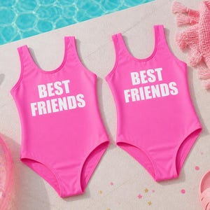 May include: Two bright pink swimsuits with the text "BEST FRIENDS" in white block letters. The swimsuits are one-piece with a scoop neck and are near a pool. The swimsuits are likely for children.