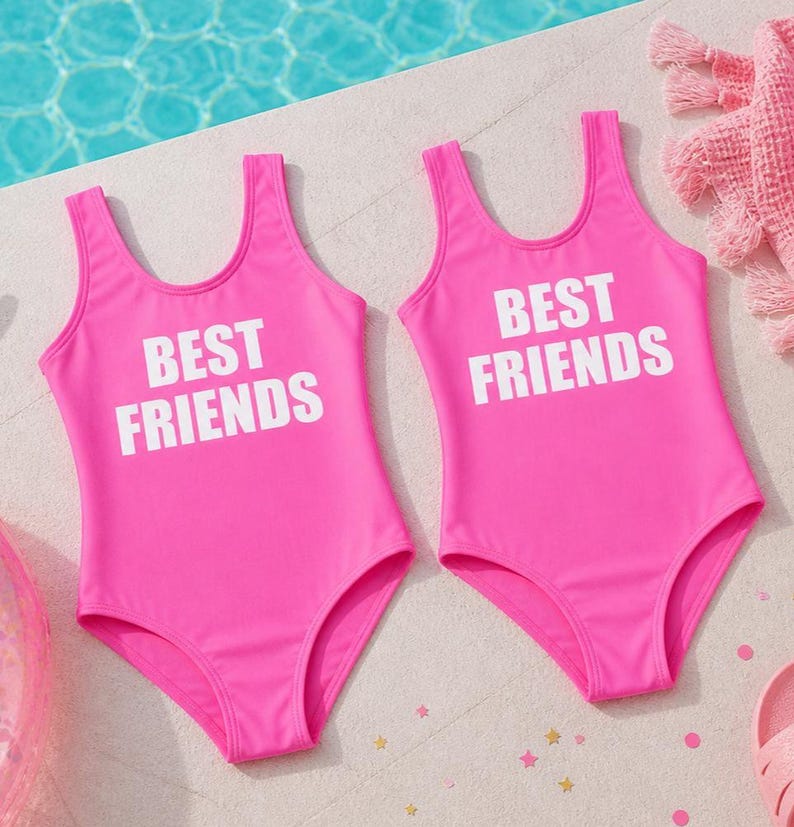 May include: Two bright pink swimsuits with the text "BEST FRIENDS" in white block letters. The swimsuits are one-piece with a scoop neck and are near a pool. The swimsuits are likely for children.