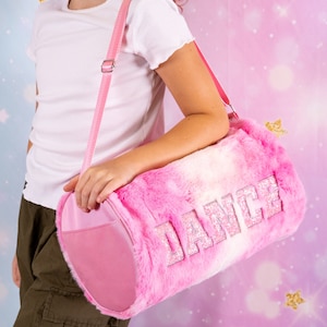 May include: A pink and white faux fur duffel bag with a glittery "DANCE" design. The bag has a pink adjustable shoulder strap.