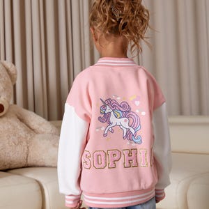 May include: Pink and white varsity jacket with a unicorn graphic and the name "SOPHIA" in gold lettering. The jacket has white sleeves and striped trim on the collar, cuffs, and hem. The unicorn is depicted with a rainbow mane and tail.