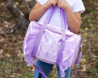 Puede incluir: Bolso de danza morado con una silueta de bailarina blanca y el nombre "Christina" bordado en la parte delantera. El bolso tiene un ribete con volantes y una correa de hombro morada.