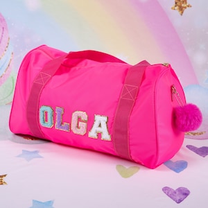May include: A bright pink duffel bag with a fluffy pink pom-pom zipper pull. The bag has the name "OLGA" spelled out in colorful, glittery letters.