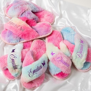 May include: Four pairs of pink and blue fuzzy slippers with criss-cross straps. Each slipper has a butterfly design and is personalized with a name: Emilia, Diana, and two more slippers with the name Emilia.