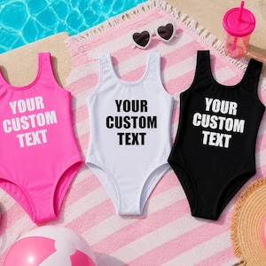 May include: Three swimsuits in pink, white, and black, each with the text "YOUR CUSTOM TEXT" in white. The swimsuits are on a pink and white striped towel, with sunglasses, a drink, a beach ball, a hat, and a floatie.