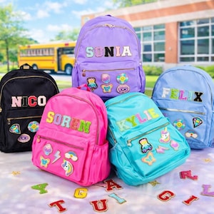 Puede incluir: Una colección de cinco mochilas en negro, morado, rosa intenso, turquesa y azul claro, cada una con parches de letras brillantes de colores que deletrean nombres como Sonia, Nico, Soren, Kylie y Felix, junto con varios parches decorativos.
