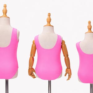 May include: Three pink swimsuits displayed on mannequins. The swimsuits have a scoop back design and are a vibrant shade of pink. The mannequins are on metal stands against a white background. The swimsuits are likely for children.