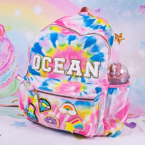 May include: A tie-dye backpack with the word "OCEAN" in gold lettering. The backpack has a front pocket with a zipper and several decorative patches. A clear water bottle with a unicorn theme is in the side pocket.