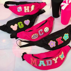 May include: A collection of fanny packs in black and hot pink. Each pack features embroidered patches, including letters, a lightning bolt, a heart, a flower, a donut, a butterfly, and a smiley face. Also, two textured tumblers, one black and one pink.