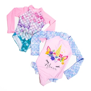 May include: Two pink long-sleeved swimsuits with mermaid scale patterns. One swimsuit has a unicorn design with the name "Alicia" and the other has the name "Noelle".
