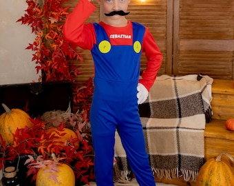 May include: A child dressed as Super Mario for Halloween. The costume is red and blue with a yellow M on the hat. The child is wearing a fake mustache and holding the hat with one hand. The costume has the name "Sebastian" written on the front.