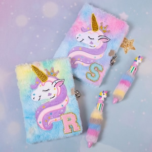 May include: Two fluffy unicorn journals with lock closures and matching pens. The journals are pastel blue, pink, and yellow with unicorn appliques, gold horns, and initial letters. The pens have unicorn toppers and colorful fur.