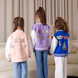 May include: Three children wearing personalized varsity jackets. The jackets are pink, purple, and blue with white sleeves. The jackets have embroidered names on the back: "Eva", "Nika", and "Sophia".