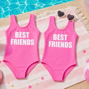 May include: Two bright pink swimsuits with the text "BEST FRIENDS" in white. The swimsuits are on a striped towel, with heart-shaped sunglasses, a beach ball, and a pink drink with a straw. The scene is set near a pool.