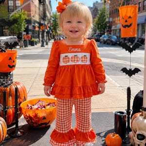 May include: A long-sleeved orange top with a white smocked panel featuring three orange pumpkins and the name Emma embroidered in white, paired with orange and white gingham bell-bottom pants with ruffled hems and an orange hair bow.