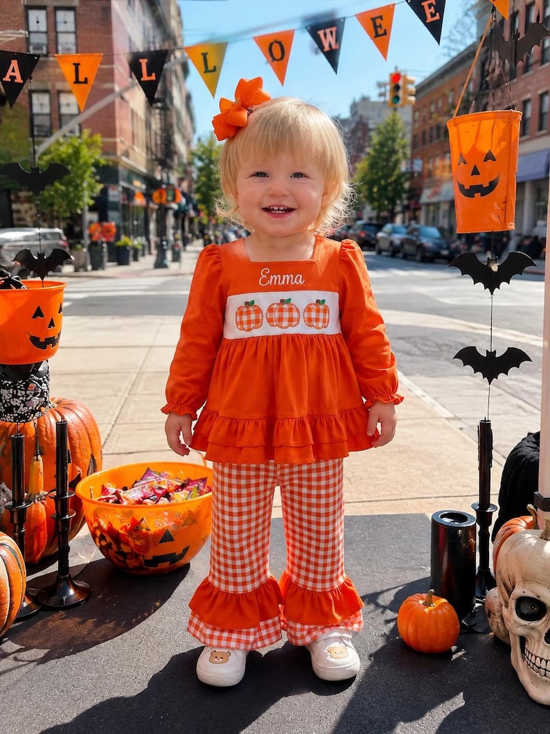 May include: A long-sleeved orange top with a white smocked panel featuring three orange pumpkins and the name Emma embroidered in white, paired with orange and white gingham bell-bottom pants with ruffled hems and an orange hair bow.