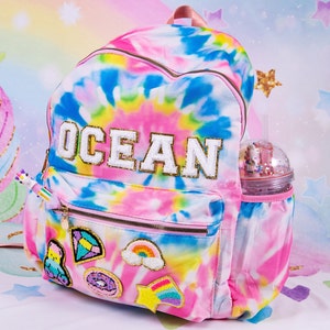 May include: A pink and blue tie-dye backpack with the word "OCEAN" in white letters with gold glitter. The backpack has a variety of colorful patches, including a rainbow, a donut, a star, and a diamond. The backpack has a side pocket with a clear plastic water bottle with a straw.