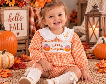 May include: An orange and white gingham long-sleeve romper with a white Peter Pan collar featuring lace trim. The chest has smocking with the embroidered text 'little pumpkin' between two pumpkins. The name 'Ava' is embroidered on the collar.