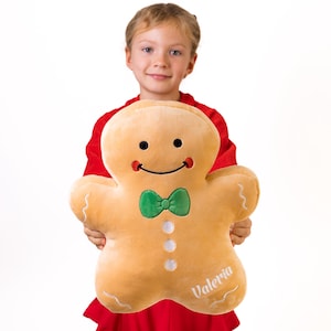 May include: A child holds a large gingerbread man plush toy. The toy is brown and has a green bow tie, white buttons, and a smiling face. The toy is personalized with the name "Valeria".
